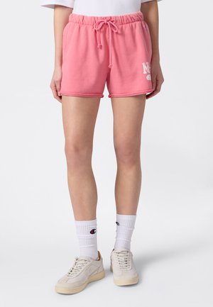 Osoba ubrana w różowe szorty ze sznurkiem, do tego białe skarpetki z logo Champion oraz kremowe trampki na jednolitym tle.