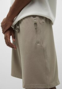 Shorts beiges en tissu texturé, avec une poche zippée sur le côté et une taille à cordon, portés avec une chemise de couleur claire.