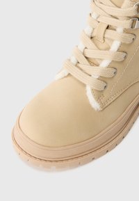 Stivale ankle in suede beige con fodera in pelliccia sintetica bianca, punta rotonda e suola piatta in gomma. Presenta lacci beige e occhielli in metallo per una vestibilità sicura.