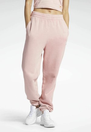 Kobieta w jasnoróżowych dresach, krótkim topie i białych trampkach stoi z rękami w kieszeniach na białym tle.