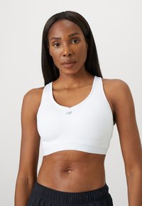 Kvinne iført en hvit sports-bh og svarte treningsshorts, stående mot en ensfarget lys bakgrunn, ser rett frem.