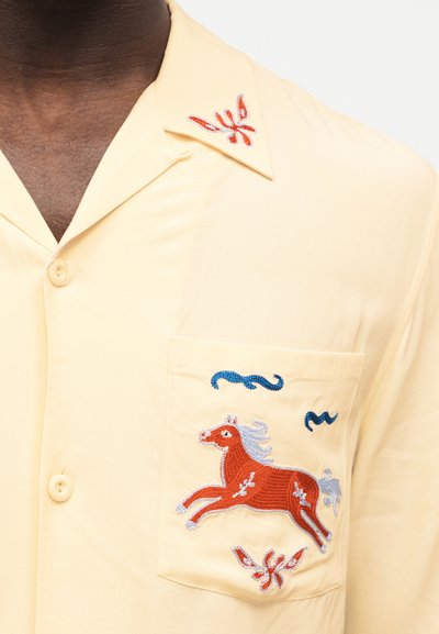 Κοντινή λήψη ενός κίτρινου πουκάμισου με κουμπιά, που διαθέτει κεντημένο κόκκινο άλογο και διακοσμητικά μπλε και κόκκινα σχέδια στην τσέπη και το γιακά.