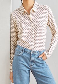 Camicia a maniche lunghe color crema con un motivo geometrico bordeaux, design con colletto e una texture morbida. Abbinata a jeans in denim azzurro chiaro.