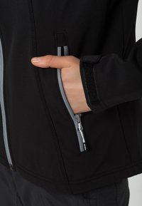 Sort softshell jakke med grå lynlåse, der har en lynlåslomme og velcro lukning ved ærmet. Glat tekstur, atletisk pasform.