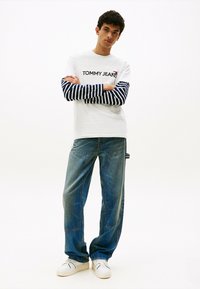 Camiseta blanca con mangas largas a rayas en azul y blanco, logo "TOMMY JEANS", combinada con jeans de mezclilla azul sueltos y zapatillas blancas.