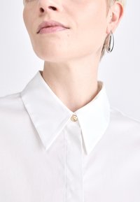 Camisa blanca de cuello con un diseño estructurado, que presenta un botón dorado brillante en el cuello. Textura suave, corte entallado y bordes nítidos.