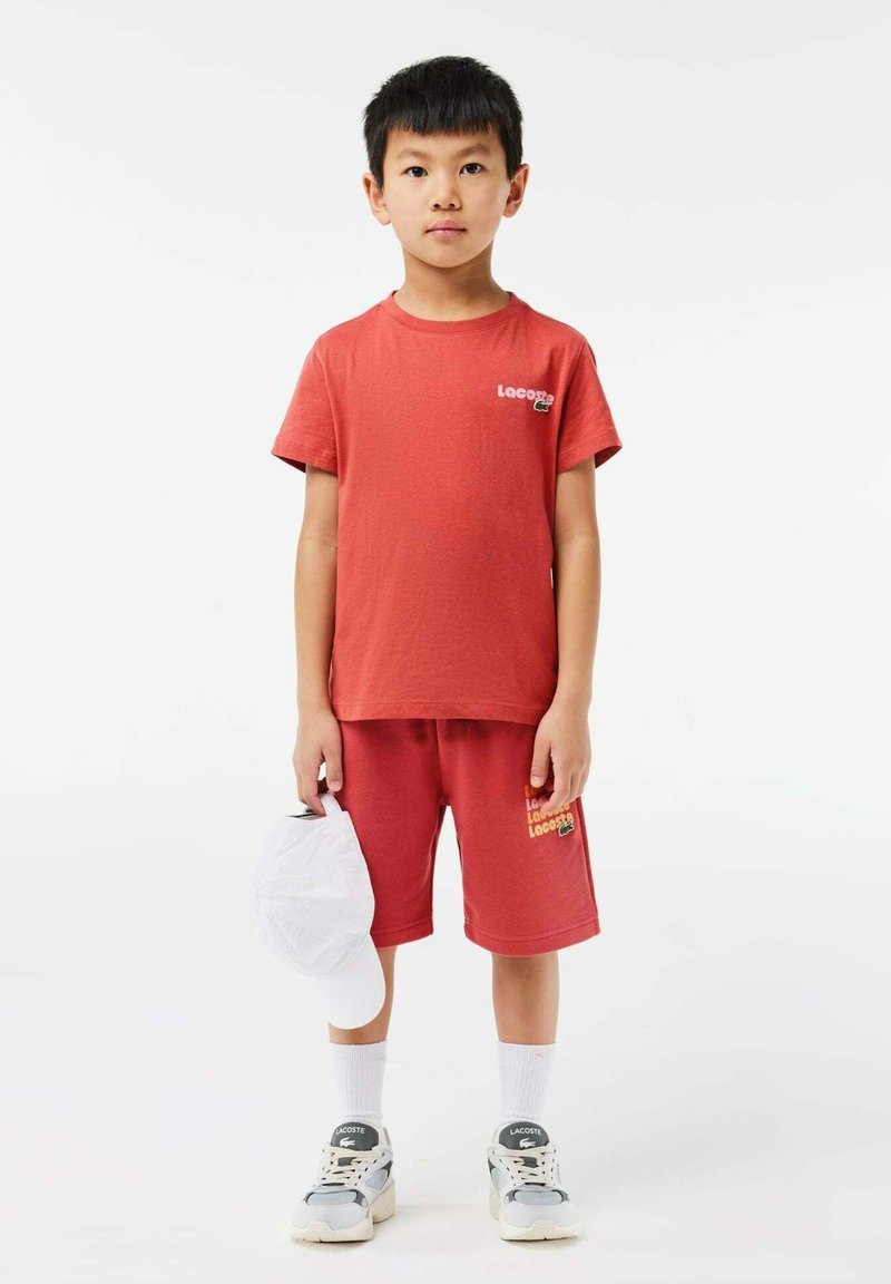 Rotes Baumwoll-T-Shirt und Shorts-Set mit Lacoste-Logo. Weiße Mütze in der Hand. Graue Sneakers mit Akzenten. Weiße Sneakersocken. Einfaches Design.