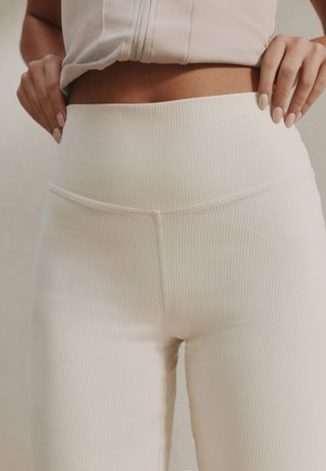 Žena upravuje si vysoký pás rebrovaných legín krémovej farby, oblečená v krátkom svetlo béžovom topíku, s rukami položenými na bokoch pri páse.