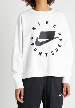 Osoba ubrana w białą bluzę Nike Sportswear z dużym czarnym logo i czarne spodnie, stojąca na jednolitym tle.
