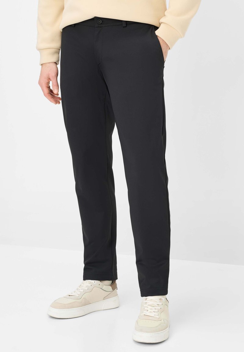 Pantalon noir sur mesure en tissu léger, avec devant plat, texture lisse et coupe ajustée, associé à des baskets claires.
