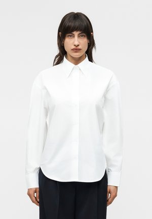 Kobieta o ciemnych włosach, ubrana w białą koszulę na guziki z długim rękawem i czarne spodnie, stojąca na tle jednolitego, jasnego tła.