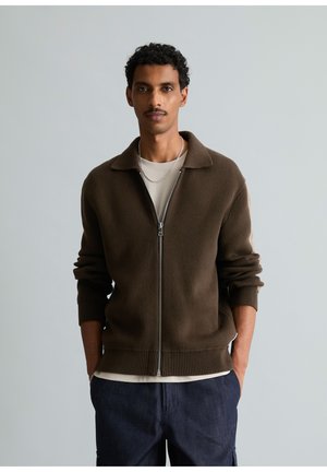 Mann mit lockigem Haar und Schnurrbart, der einen braunen Zip-Up-Pullover über einem hellen Hemd und dunklen Hosen trägt und mit den Händen in den Taschen steht.