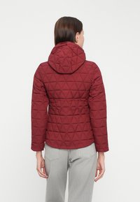 Veste matelassée rouge avec capuche, manches longues et motif triangulaire, associée à un pantalon gris clair, vue de dos.