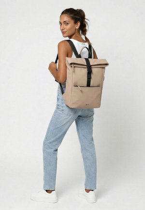 Vrouw met paardenstaart, die lichtblauwe spijkerbroek, witte sneakers, wit mouwloos topje en beige rugzak met zwarte bandjes draagt, staand zijwaarts.