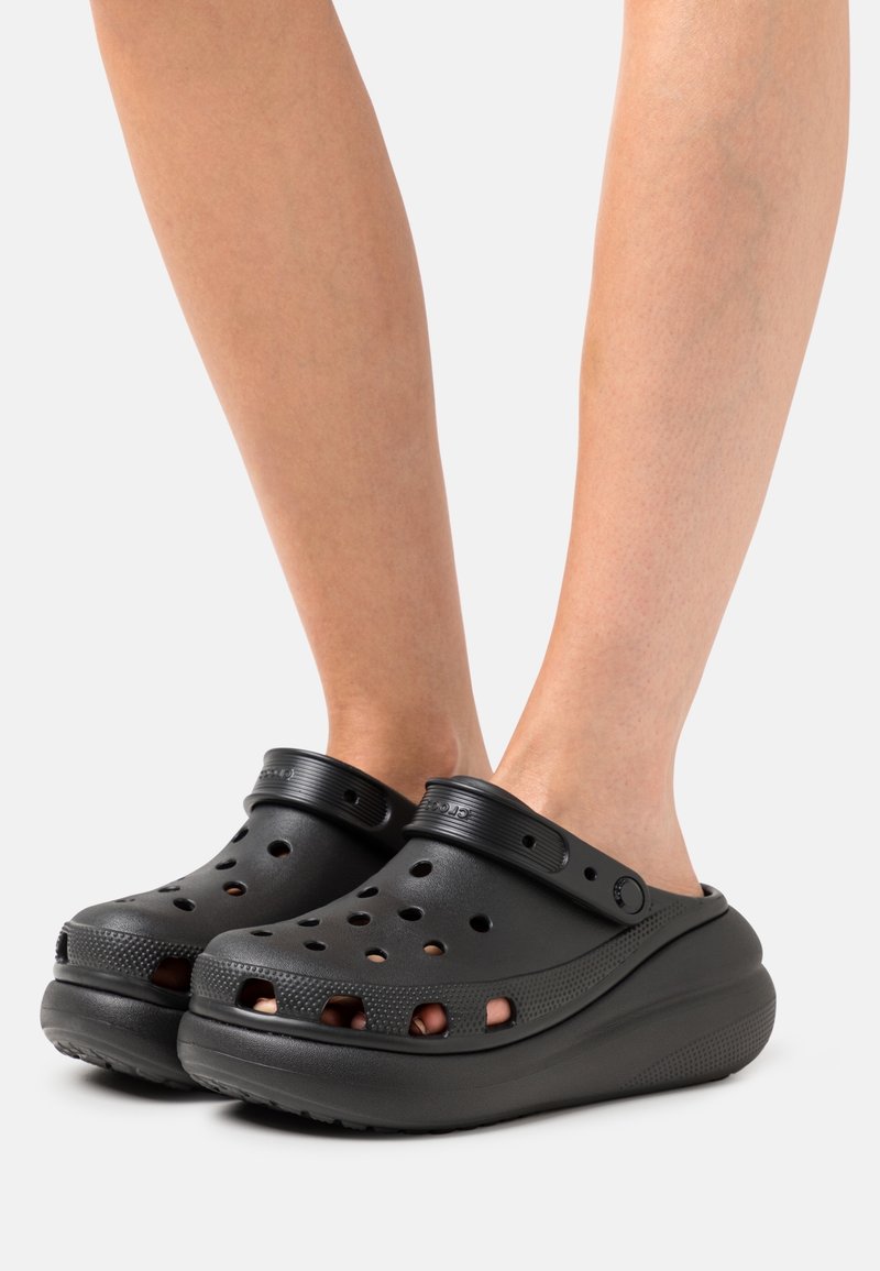 Pieds portant des sabots noirs à enfiler avec des trous de ventilation et des semelles épaisses et surélevées sur un fond clair uni.