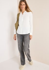 Chemise blanche à boutons, jeans gris fuselés, baskets crème avec des accents texturés. Le mannequin tient un sac vert olive, debout devant un fond neutre.