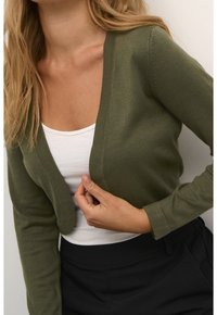 Cardigan en tricot vert olive avec un col en V profond et des manches longues, porté par-dessus un haut blanc, présentant une silhouette ajustée.