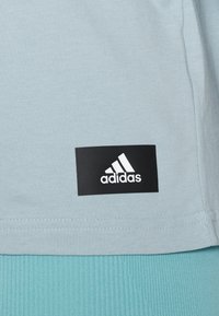 Světle modrá látka s černou obdélníkovou nášivkou loga Adidas přišitou blízko lemu nad tyrkysovou žebrovanou látkou.