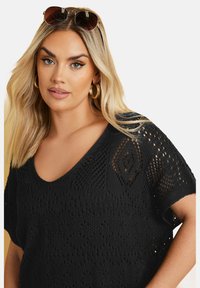 Top negro de punto con corte suelto, que presenta un escote en V, un patrón abierto y mangas cortas. Diseño texturizado con formas de diamante y acentos de celosía.