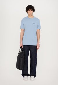 Tricou scurt cu mânecă scurtă de culoare albastru deschis, cu un logo mic închris, asortat cu blugi largi de culoare albastru închis și o geantă tote neagră. Adidași albi finalizează look-ul.