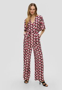 Geprinte jumpsuit met een V-hals, korte mouwen, wijde benen en een strik ceintuur. Kleuren omvatten roze, zwart en crème met een zigzagpatroon.