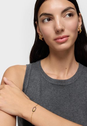 Kobieta o ciemnych włosach w szarym topie bez rękawów nosi złote kolczyki-koła oraz delikatną złotą bransoletkę z czarno-złotym owalnym zawieszkiem.