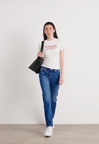 Ženska samozavestno hodi v beli majici "Tommy Jeans" in modrih kavbojkah, nosi črn torbico, proti svetli ozadju.