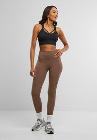 Črn športni nedrček s prečnimi naramnicami, v kombinaciji s srednje visokimi rjavimi leggins. Model nosi sive športne copate in bele ankel nogavice.
