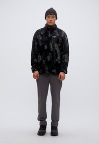 Μαύρη και γκρι μπλούζα από φλις με σχέδια, ψηλός γιακάς, τσέπη με φερμουάρ στο πλάι. Συνδυασμένη με γκρι παντελόνι και μαύρα ορειβατικά μποτάκια, ολοκληρωμένη με μαύρο σκουφάκι.