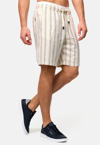 Shorts beiges rayés avec un cordon de serrage à la taille, présentant des lignes verticales sombres et des poches latérales. Associés à des baskets navy.
