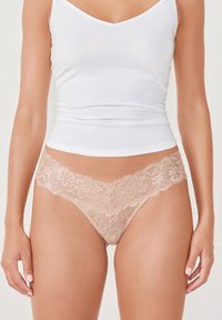 Spitzen-Thong in sanftem Beige mit floralem Muster am Bund. Kombiniert mit einem weißen, figurbetonten Tanktop. Glatte Textur und zartes Design.