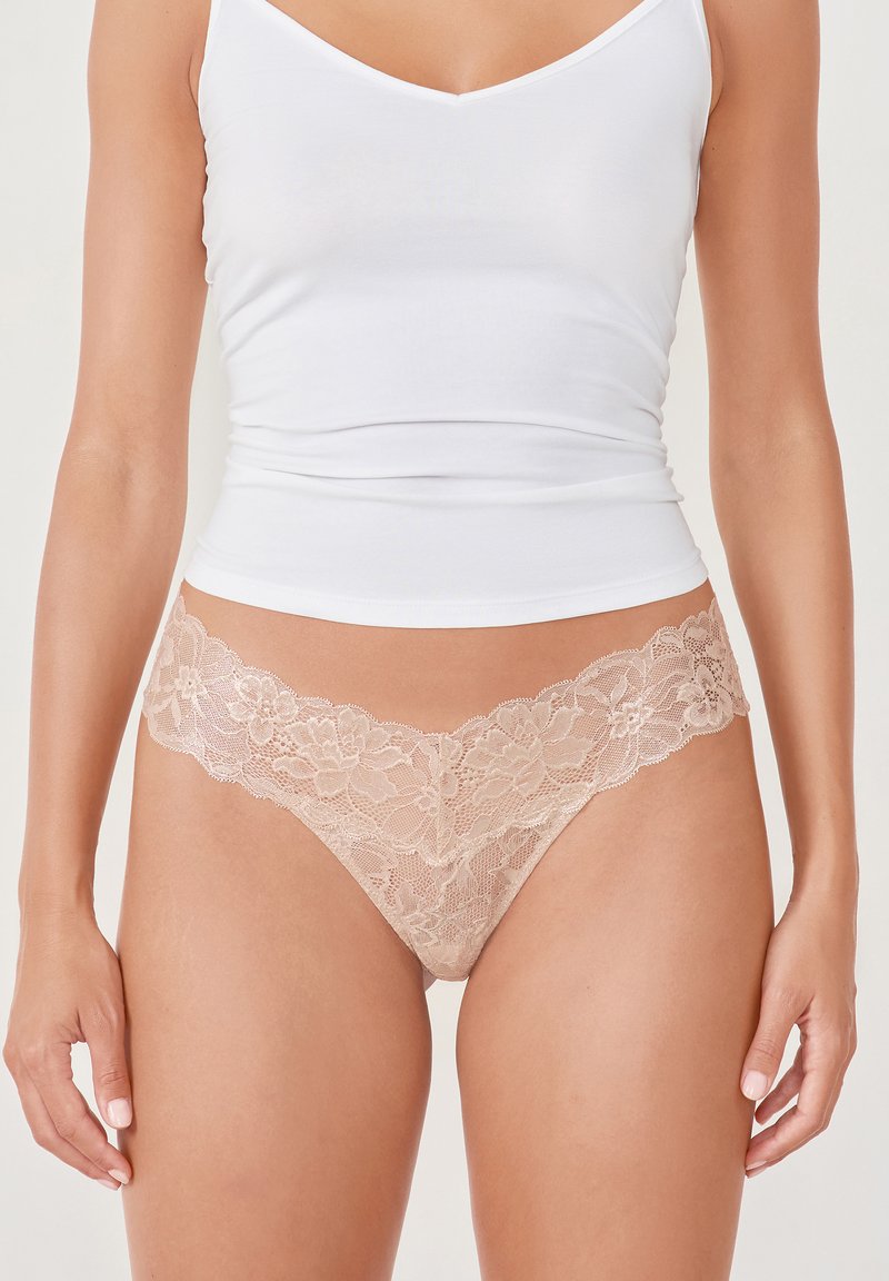 Spitzen-Thong in sanftem Beige mit floralem Muster am Bund. Kombiniert mit einem weißen, figurbetonten Tanktop. Glatte Textur und zartes Design.