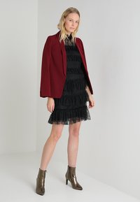 Burgunderfarget blazer over en svart spetssløyd kjole med lag, som har volanger og en strukturert finish, kombinert med metalliske ankelstøvletter.