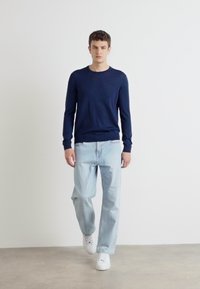 Tengerészkék kerek nyakú pulóver, világoskék, laza szabású farmernadrág és fehér sportcipők. Az anyag lágy, sima tapintású. Minimalista stílus.