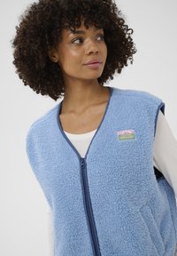 Frau mit lockigem Haar, die eine hellblaue Fleece-Weste mit Reißverschluss und einem kleinen Aufnäher über einem weißen Langarmshirt trägt und zur Seite blickt.