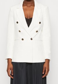Blazer bianco doppiopetto con rever, bottoni dorati e due tasche laterali, abbinato a un top nero e a una gonna plissettata.