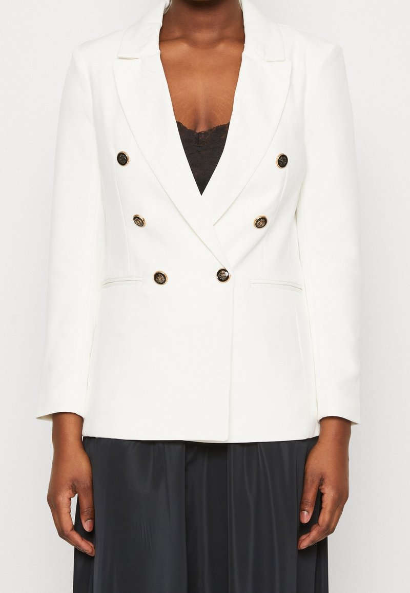 Blazer bianco doppiopetto con rever, bottoni dorati e due tasche laterali, abbinato a un top nero e a una gonna plissettata.