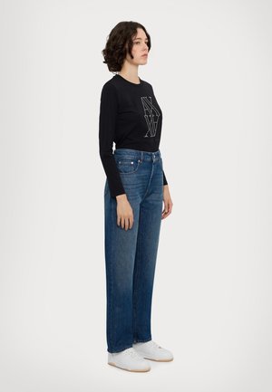 Žena s krátkymi kučeravými vlasmi, oblečená v čiernej dlhé rukávy, modrých širokých džínsach a bielych teniskách, stojaca bokom na jednoduchom pozadí.