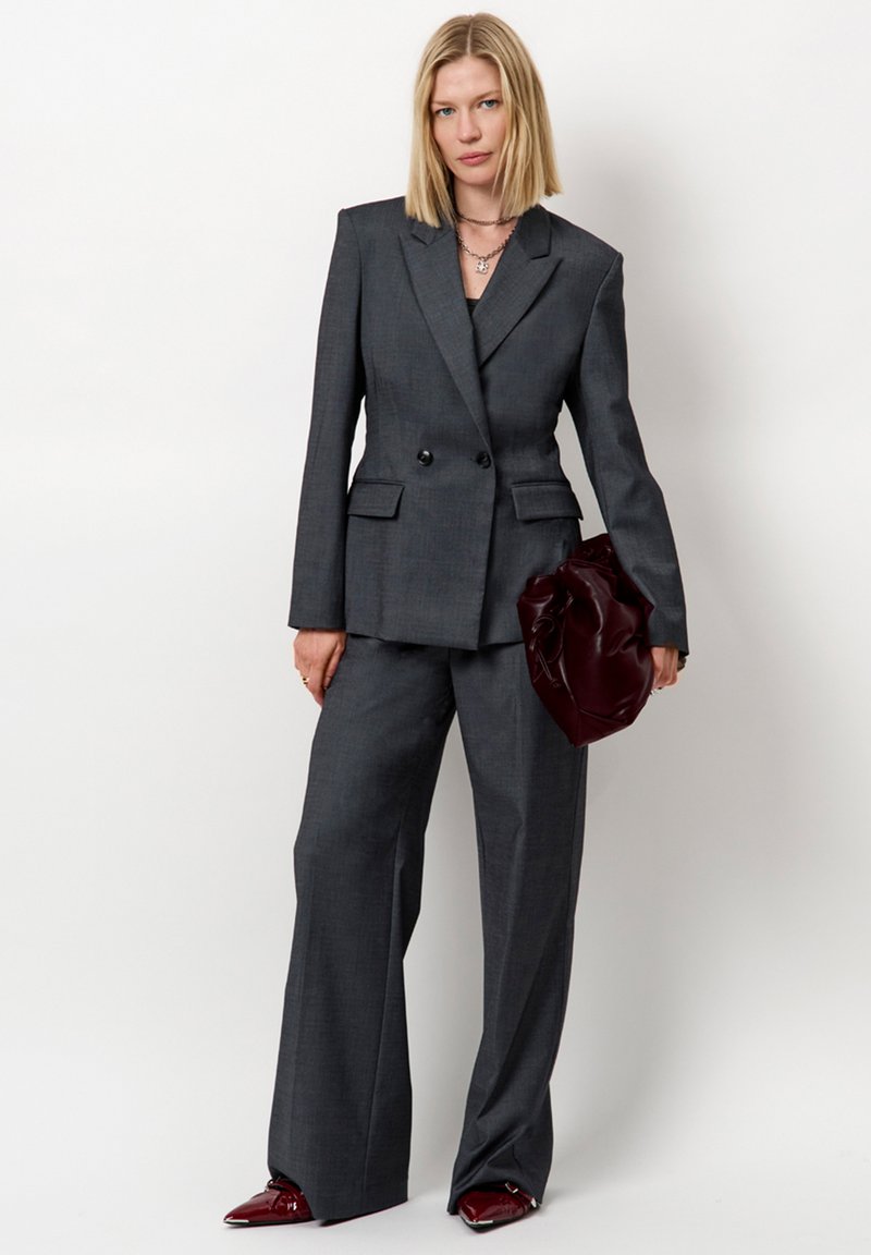 Vrouw in een op maat gemaakt donkergrijs pak met een wijde broek, die een donker bordeaux kleurige slouchy clutch vasthoudt, staand tegen een witte achtergrond.