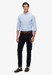 Lyseblå stribet skjorte med knapper, mørke jeans, brun flettet bælte og brune loafers. Modellen står lige med hænderne afslappet ned langs siden.