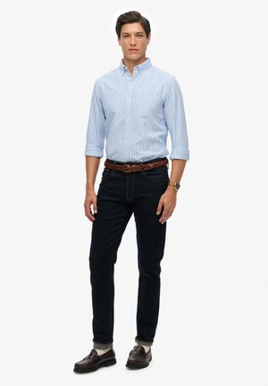 Helles, blau gestreiftes Hemd mit Knopfleiste, dunkle Jeans, brauer geflochtener Gürtel und braune Slipper. Das Model steht aufrecht mit entspannten Händen an den Seiten.