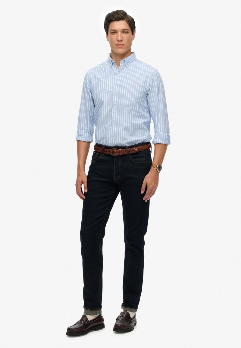 Lyseblå stribet skjorte med knapper, mørke jeans, brun flettet bælte og brune loafers. Modellen står lige med hænderne afslappet ned langs siden.