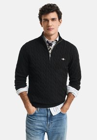 Young man with dark curly hair wearing a black cable-knit half-zip sweater, striped shirt with rolled sleeves, and blue jeans with hands in pockets.