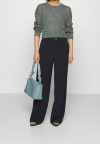 Kvinna iförd grön stickad tröja och svarta vida byxor, som håller en ljusblå vävd handväska med knutna handtag.