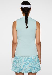 Kobieta z warkoczami, ubrana w bezrękawnik w jasnoniebieskim kolorze, wzorzystą niebieską spódnicę, białą daszek i rękawiczkę golfową, zwrócona tyłem na jednolitym tle.