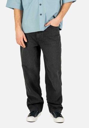 Hombre con vaqueros negros holgados, zapatillas negras y blancas, y una camisa azul clara abotonada, de pie con una mano en el bolsillo.