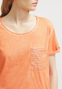 Žena v měkkém oranžovém tričku s krátkým rukávem, které má vlevo kapsu pokrytou flitry, se dívá doprava.