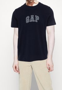 Námořnická modrá bavlněná tričko s šedou vyšívanou logem "GAP". Krátké rukávy a kulatý výstřih. V kombinaci se světlými tanovými manšestrovými kalhotami.
