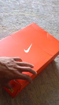 Caja de zapatos Nike en un brillante color naranja con un logo blanco; mano extendiéndose hacia ella. La caja presenta una textura suave y forma rectangular.