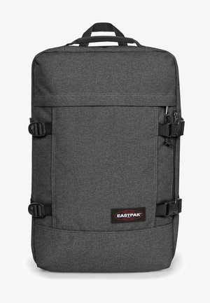 Mochila de tela gris con forma rectangular, hebillas ajustables, asa superior y un logo de marca visible. Textura suave, diseño minimalista.